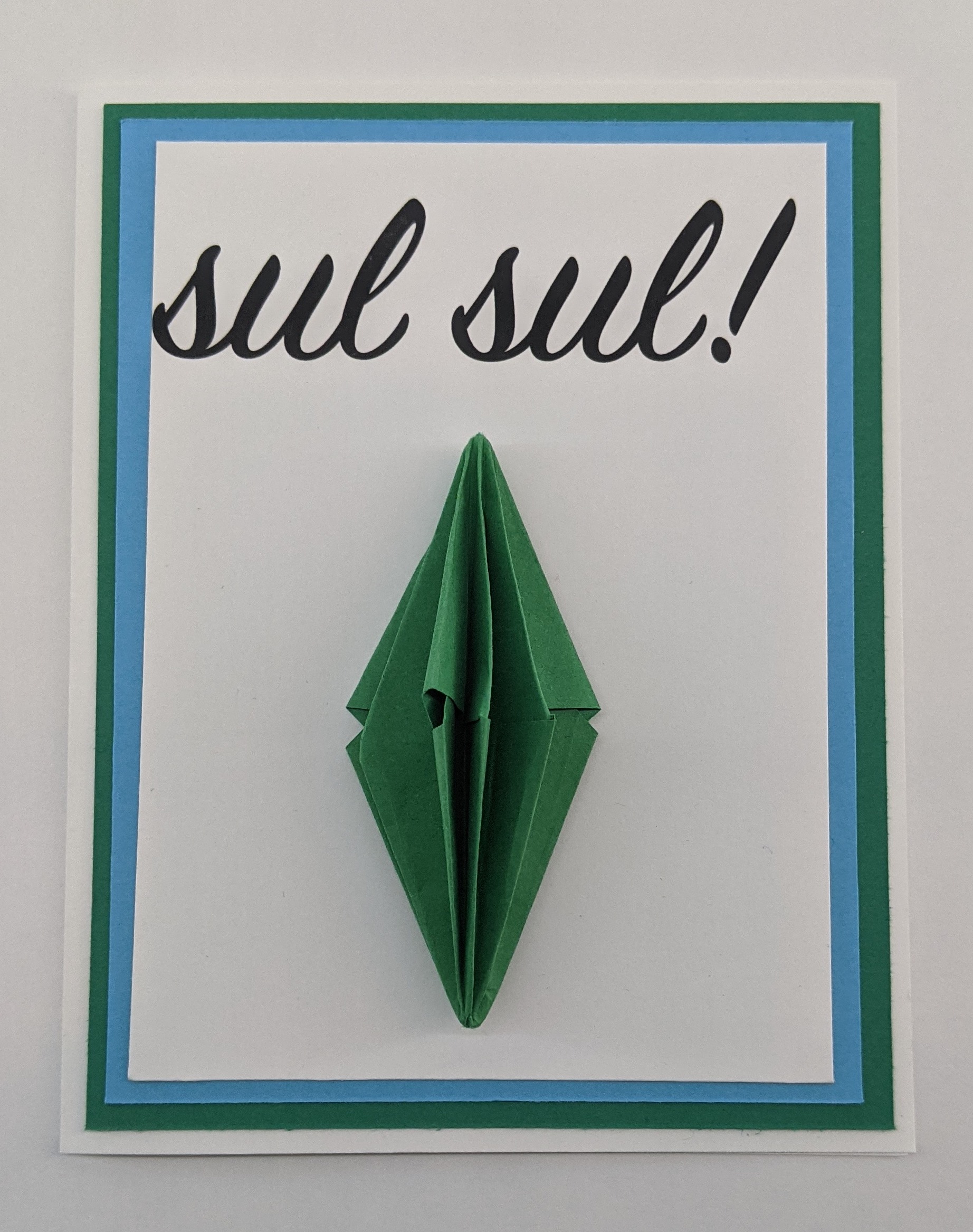 card of white paper, with blue and green accents, and a green plumbob origami and the words "sul sul" in script